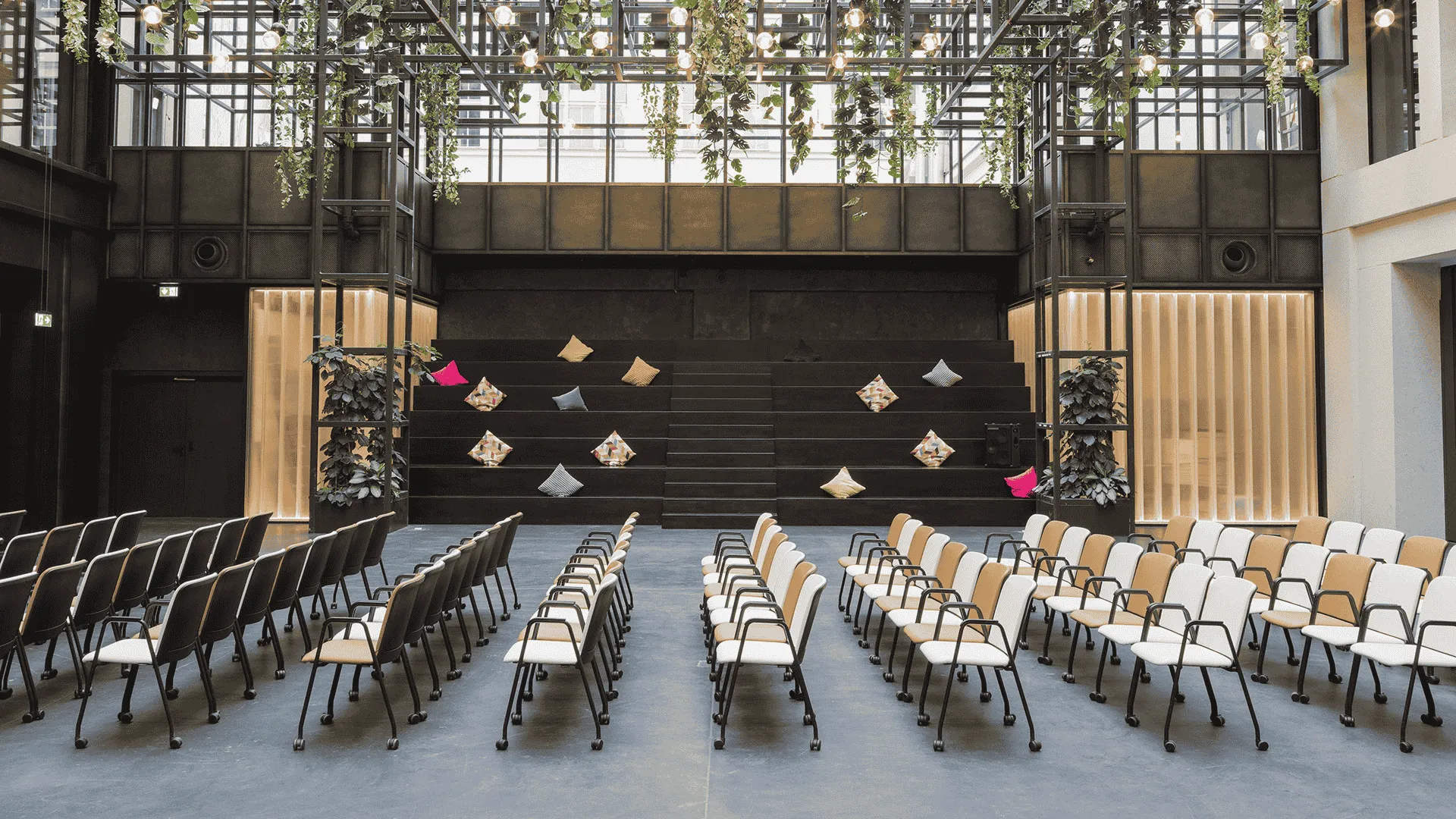 Moderne Veranstaltungshalle mit Reihen von schwarzen und weißen Stühlen, gerichtet auf eine Bühne mit hängenden Kunstinstallationen und grüner Pflanzen-Deko im oberen Bereich.