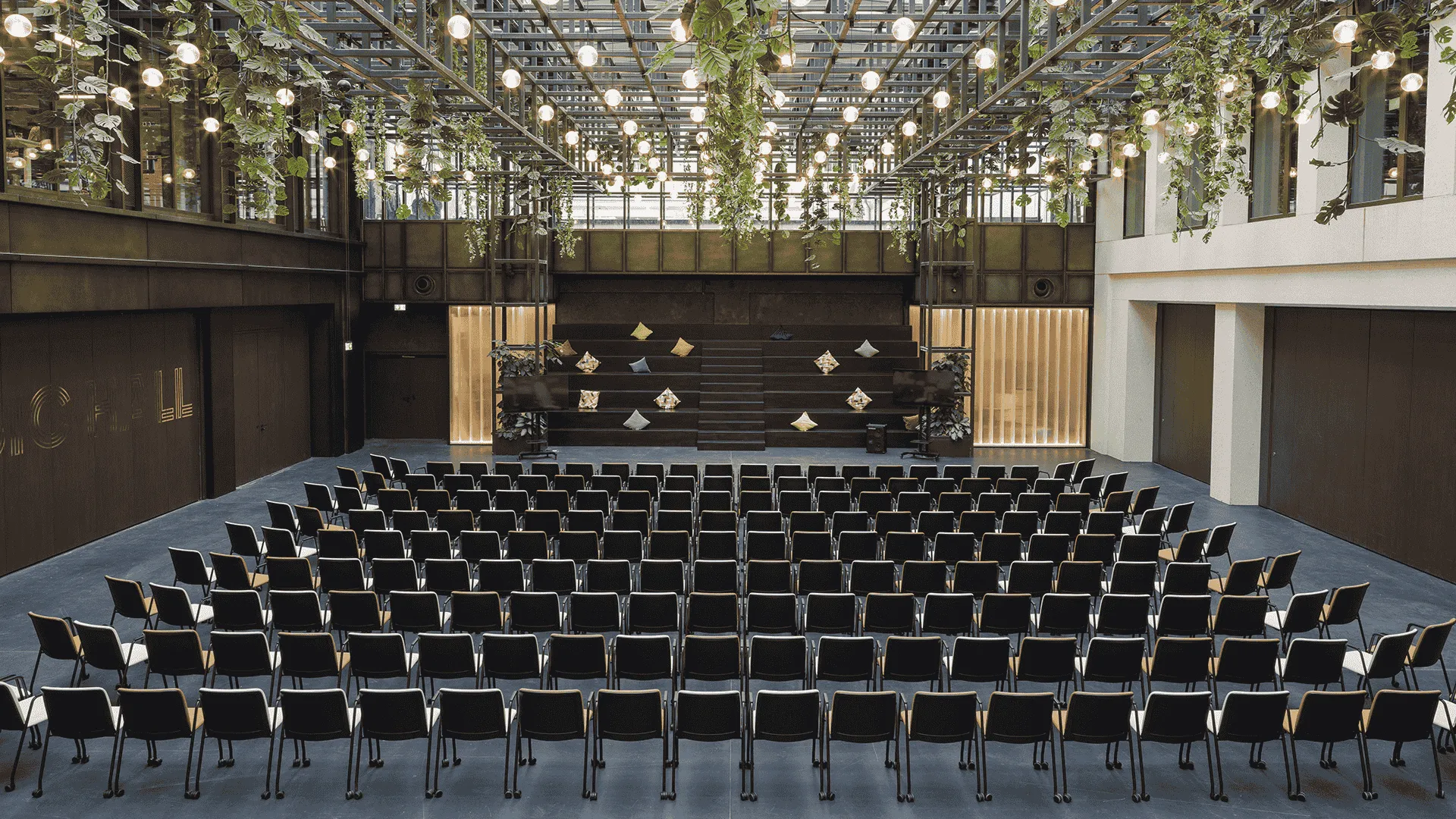 Moderne Aufführungshalle mit halbkreisförmiger Sitzanordnung aus schwarzen Stühlen, Bühne vorne, überdacht von einem Glasdach mit hängenden Pflanzen und Lichtern.