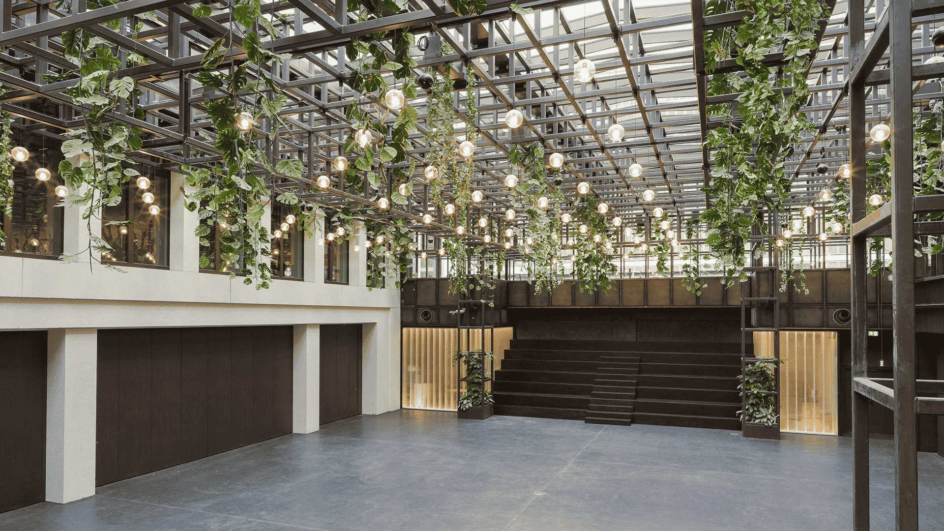 Moderne Halle mit Glasdach, beleuchtet durch zahlreiche hängende Glühbirnen, umgeben von üppigen grünen Pflanzen und einer Galerieebene aus Holz.