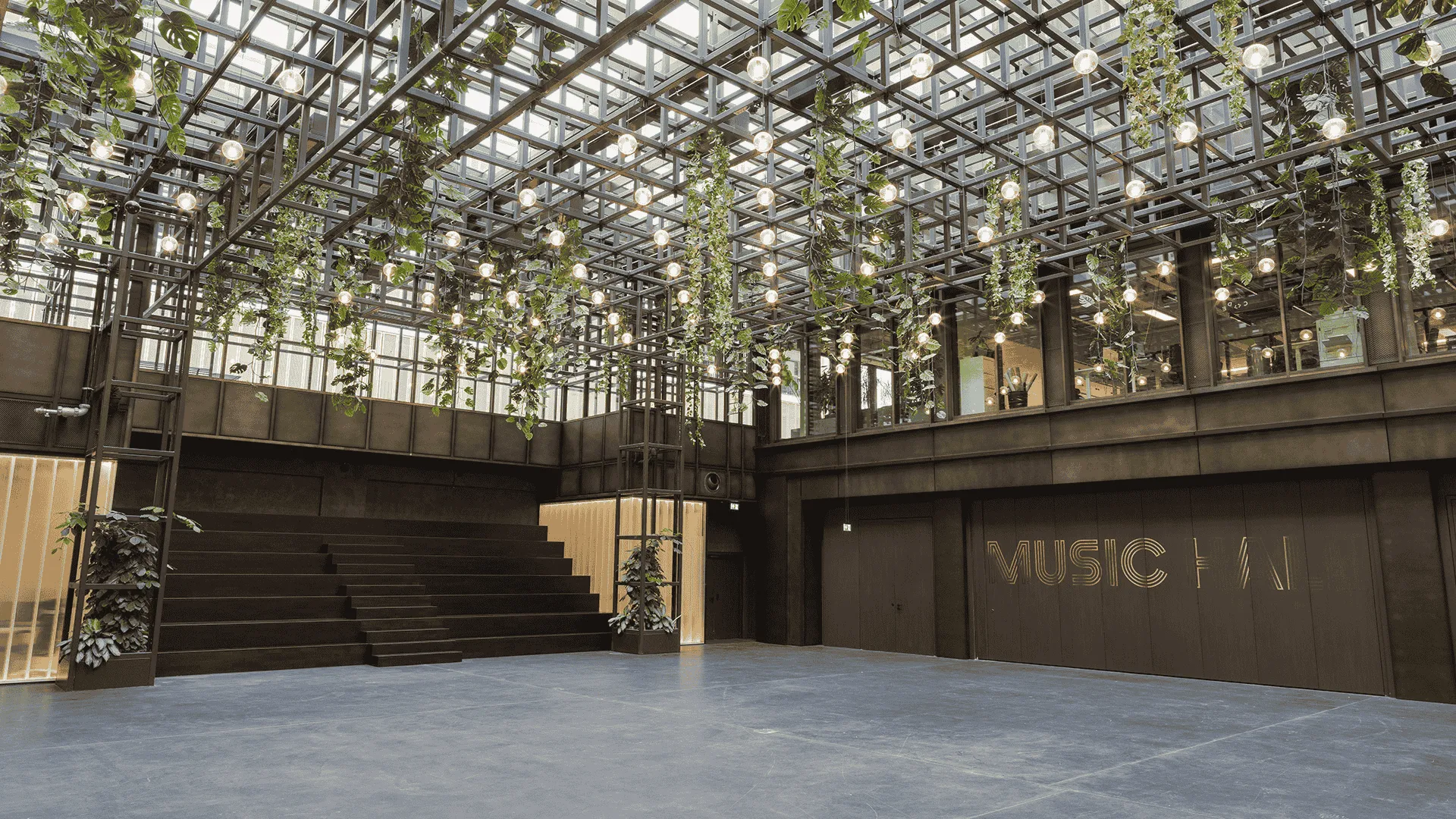 Moderne Halle mit gläsernem Dach, durch das Pflanzen hängen, beleuchtet durch zahlreiche Glühbirnen, mit einer großen Treppe und der Aufschrift 'WERK STATT' an einer Wand.